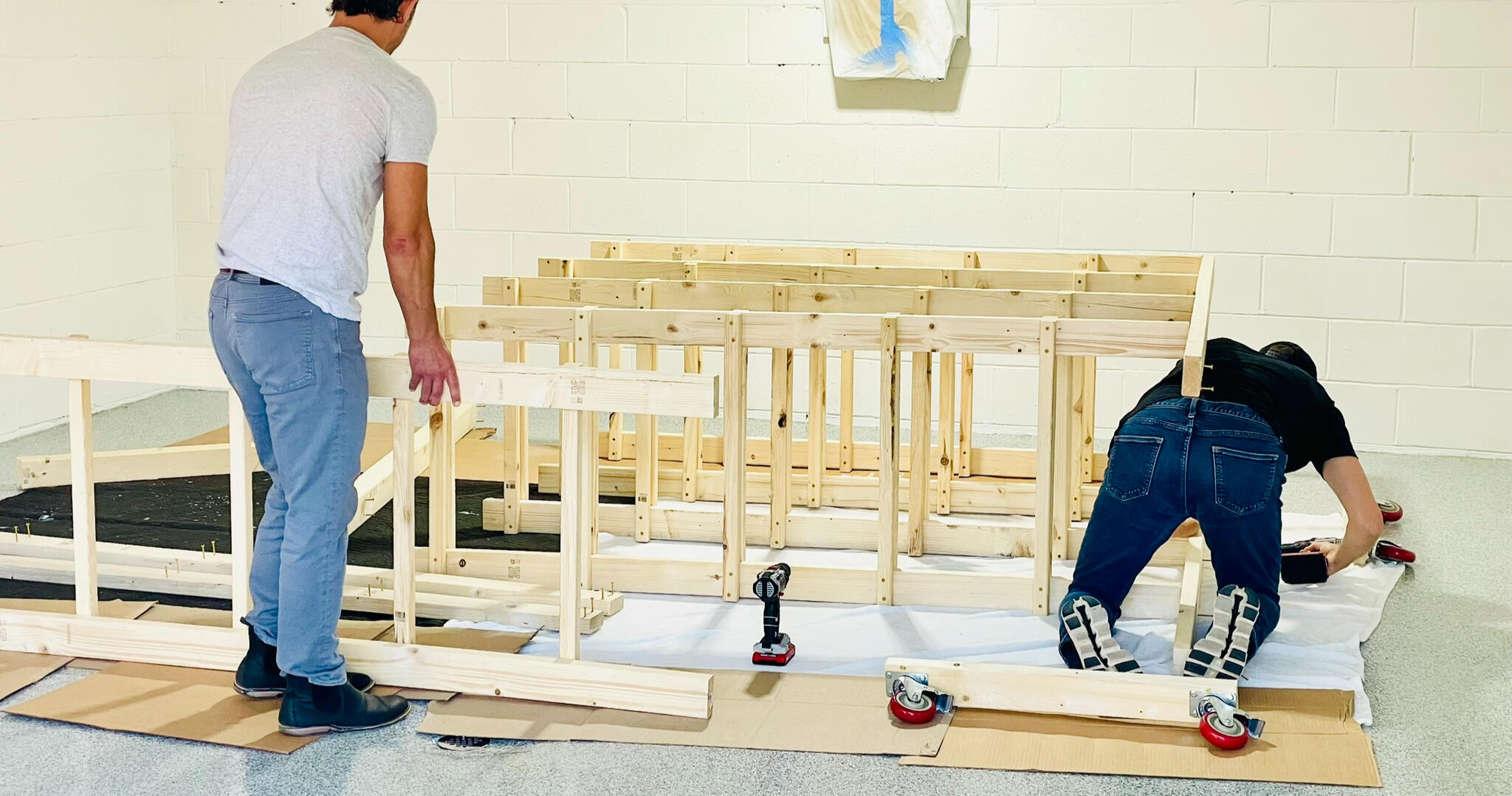 Unit Assembly Two mean are featured putting together a custom wooden storage unit. One is kneeling on the floor and screwing pieces together while the other man supports one of the wooden columns.